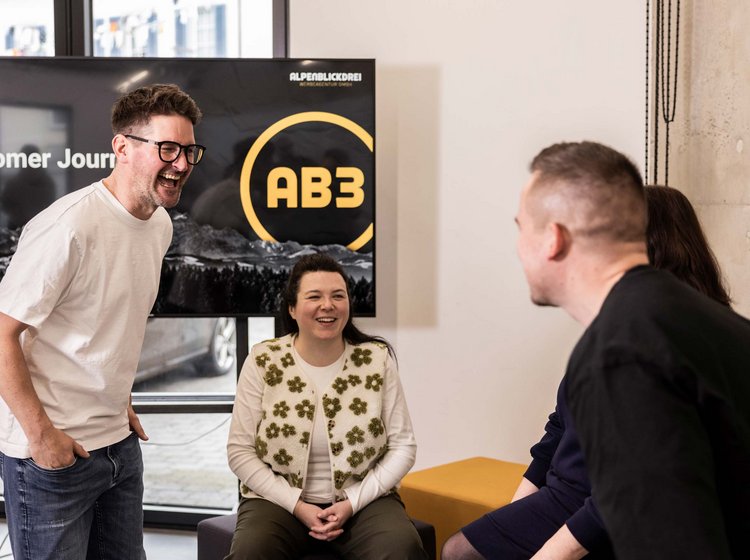 Vier Menschen in einem Gespräch, lachen und wirken freundlich in einem modernen Besprechungsraum mit einem Bildschirm im Hintergrund.