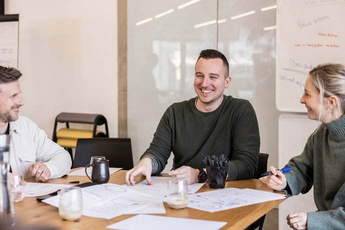Drei Personen sitzen an einem Tisch, diskutieren und lächeln während eines Meetings. Auf dem Tisch liegen Papiere mit Skizzen, und im Hintergrund ist ein Whiteboard mit handschriftlichen Notizen zu sehen.