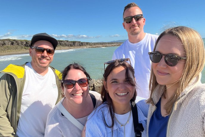 Ein Teil des AB3-Teams posiert fröhlich vor der Küste, mit strahlendem Sonnenschein und der Weite des Meeres im Hintergrund. 