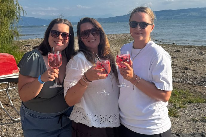 Drei AB3-Mitarbeiterinnen stoßen bei sonnigem Wetter am Ufer des Bodensees mit Drinks an.