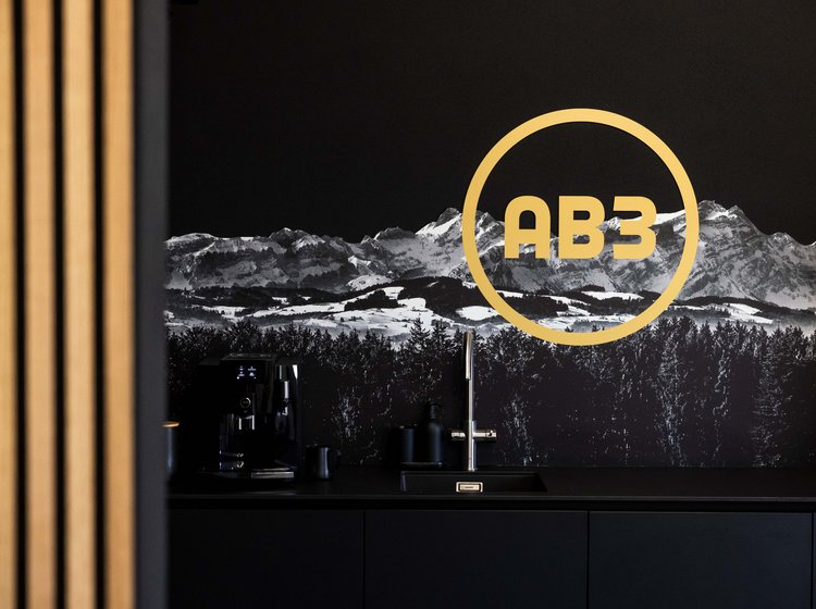 Ein moderner Küchenbereich mit schwarzer Wand und dekorativem Hintergrundbild von Bergen. Auf der Wand ein gelb beleuchtetes rundes Logo mit der Aufschrift „AB3“ in Bad Waldsee. 