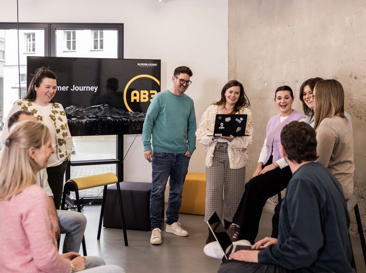 Eine Gruppe von acht jungen Menschen steht und sitzt in einem modernen, hellen Raum und diskutiert lächelnd zusammen. Im Hintergrund ist ein Bildschirm mit dem Text „Customer Journey“ zu sehen. Die Atmosphäre wirkt locker und kollaborativ.