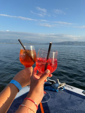 Zwei Aperitif-Gläser werden vor dem Bodensee in der Abendsonne aneinandergestoßen.