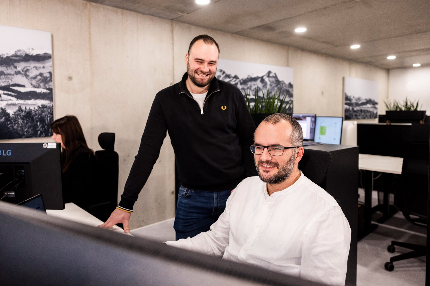 Zwei Männer der Alpenblickdrei Werbeagentur im Gespräch am Arbeitsplatz, einer steht, der andere sitzt.