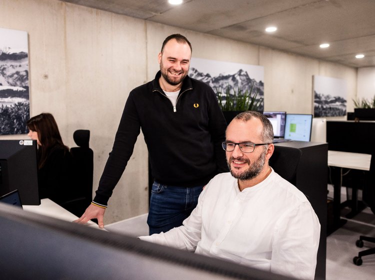 Zwei Männer der Alpenblickdrei Werbeagentur im Gespräch am Arbeitsplatz, einer steht, der andere sitzt.