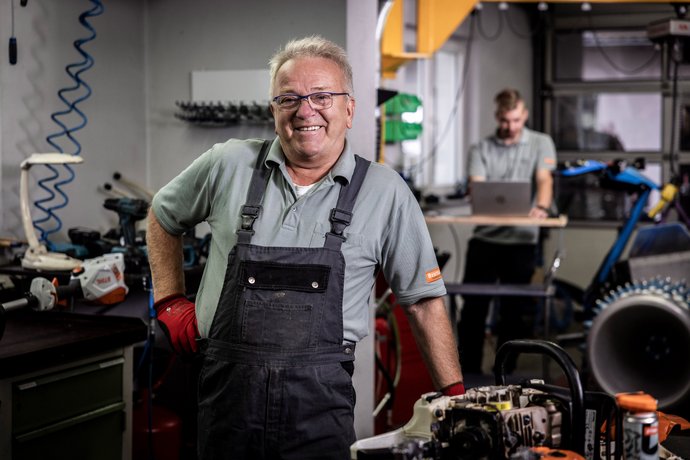 Ein älterer Mann in Arbeitskleidung steht lächelnd in einer Werkstatt mit Maschinen und Werkzeugen, im Hintergrund arbeitet ein jüngerer Mann an einem Laptop.