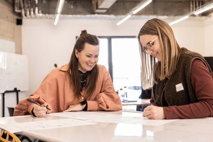 Zwei Frauen sitzen an einem Tisch und arbeiten gemeinsam an Papieren, sie lächeln und wirken konzentriert und zufrieden.