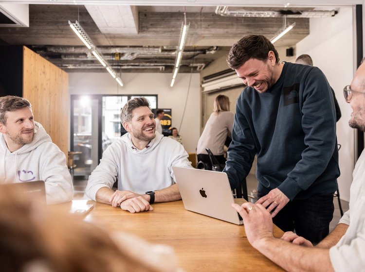 Mehrere Männer sitzen und stehen um einen Tisch, lachen und diskutieren gemeinsam. Einer zeigt etwas auf einem Laptop, während die anderen aufmerksam und freundlich reagieren.