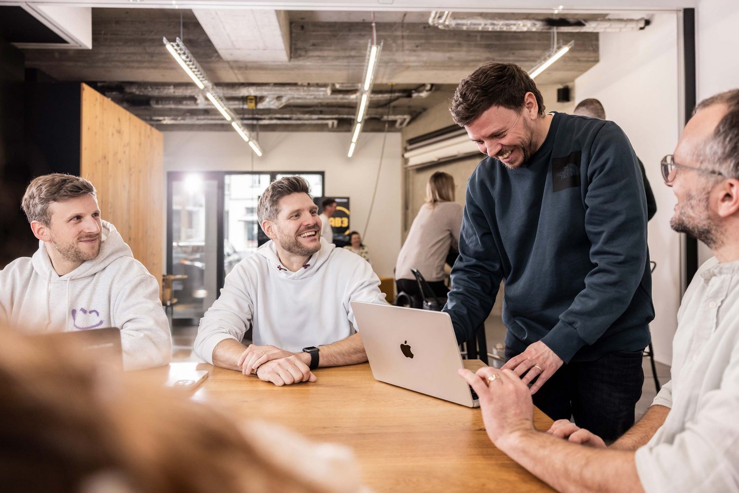 Mehrere Männer sitzen und stehen um einen Tisch, lachen und diskutieren gemeinsam. Einer zeigt etwas auf einem Laptop, während die anderen aufmerksam und freundlich reagieren.
