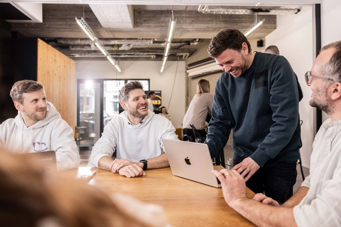 Mehrere Männer sitzen und stehen um einen Tisch, lachen und diskutieren gemeinsam. Einer zeigt etwas auf einem Laptop, während die anderen aufmerksam und freundlich reagieren.