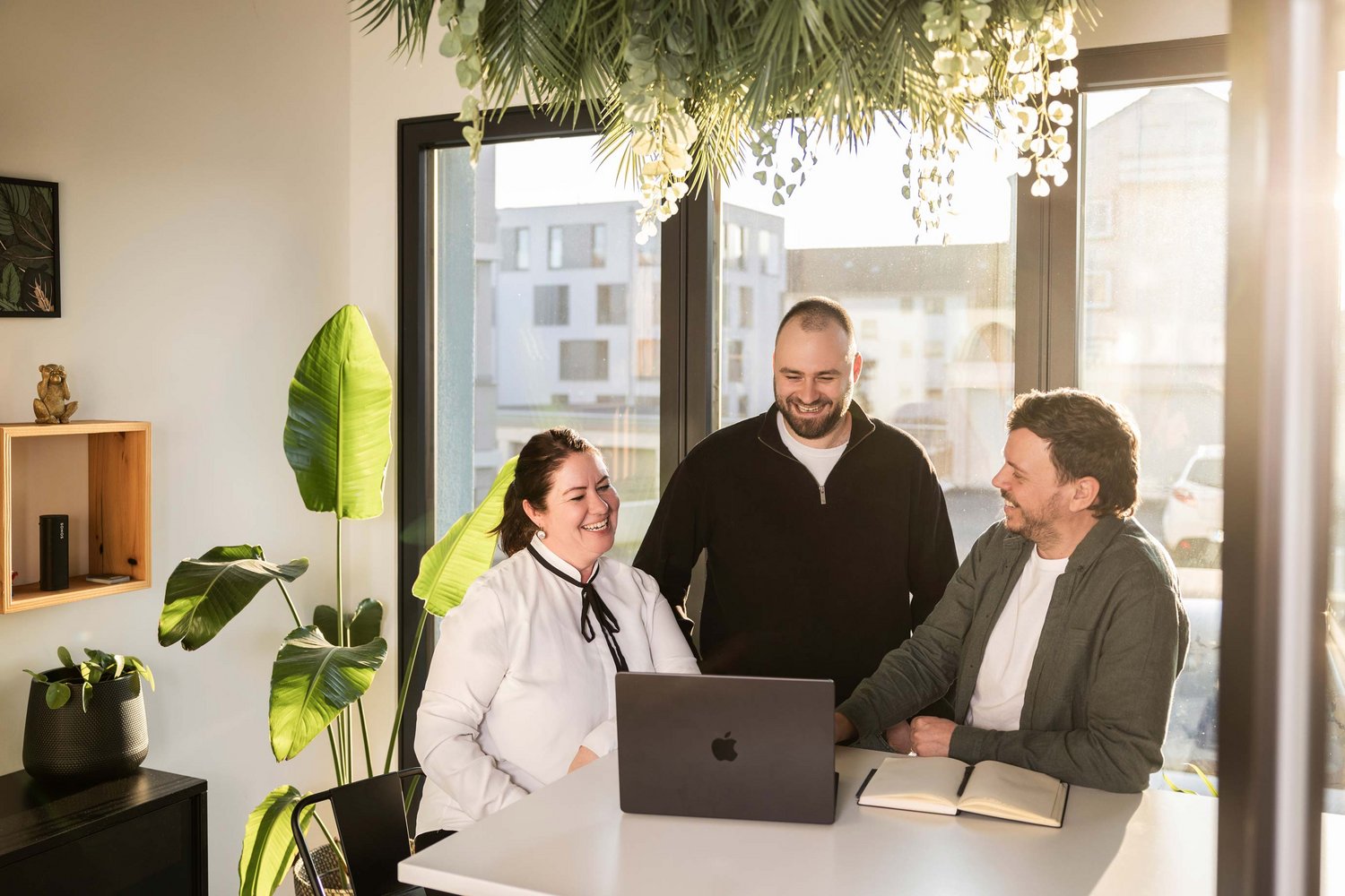 Drei Personen sitzen an einem Tisch mit einem Laptop und einem Notizbuch, lachen und führen ein entspanntes Gespräch in einem hellen, modern eingerichteten Büro mit großen Fenstern und Pflanzen.