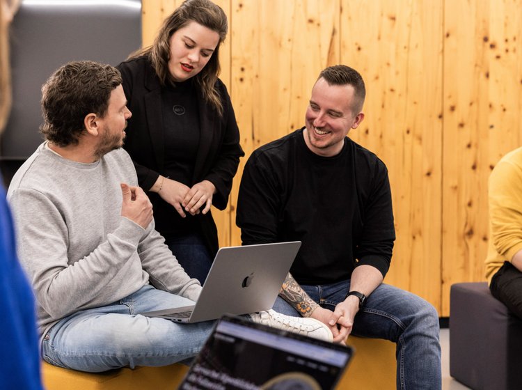 Drei Teammitglieder der Alpenblickdrei Werbeagentur im Austausch auf einer grauen Couch vor Holzdekor.