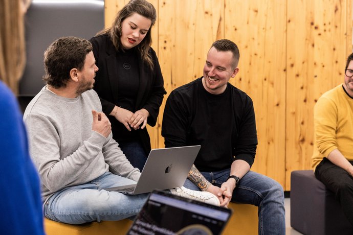 Kreativgespräch auf der Couch Drei Teammitglieder der Alpenblickdrei Werbeagentur im Austausch auf einer grauen Couch vor Holzdekor.