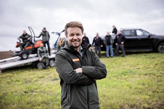 Ein lächelnder Mann steht mit verschränkten Armen auf einer Wiese, im Hintergrund mehrere Personen, Fahrzeuge und ein Anhänger mit Arbeitsgeräten. Alle tragen Outdoor-Kleidung, was auf ein Team in einem Arbeits- oder Außeneinsatz hinweist.