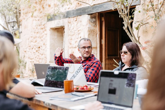 Mann mit Cap arbeitet an Laptop an einem Holztisch im Freien, daneben Snacks und Getränke, weitere Personen im Hintergrund vor Steinmauer. 