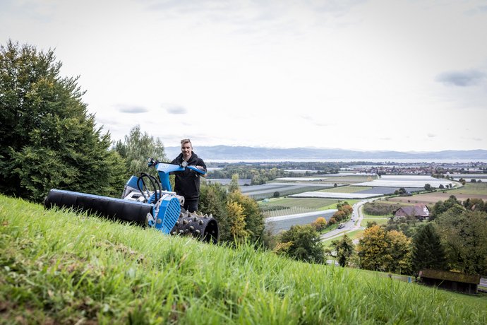 Ein Mann bedient auf einer hügeligen Wiese eine landwirtschaftliche Maschine, im Hintergrund ist eine weite Landschaft mit Feldern, Bäumen und einem See zu sehen.