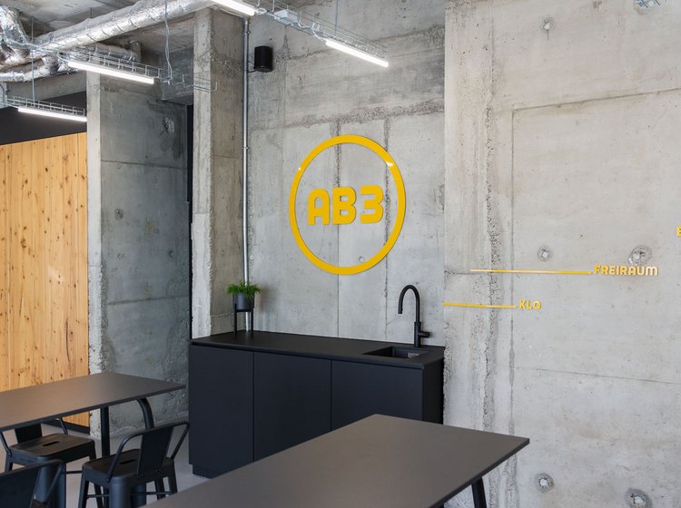 Gelbes AB3-Logo auf Betonwand vor einem modernen Tisch mit Stühlen.