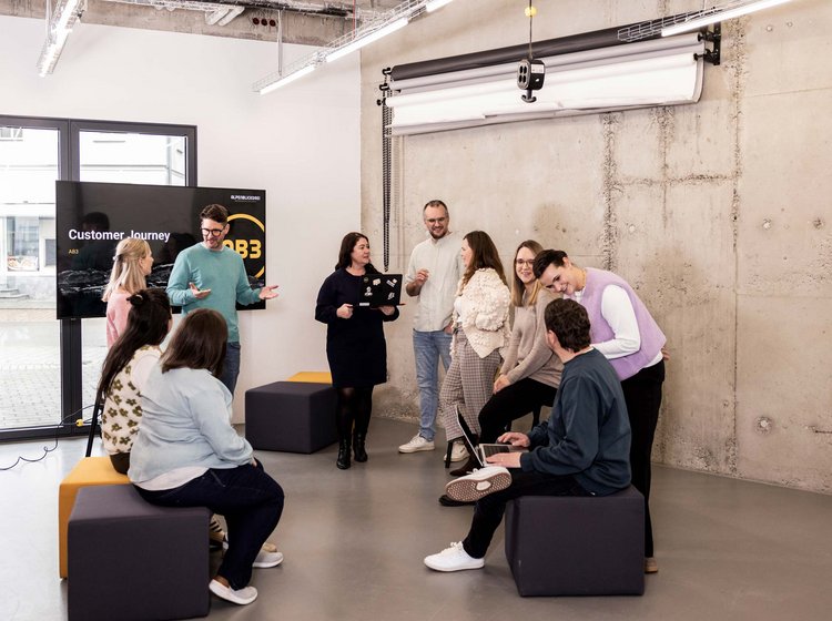 Eine Gruppe von acht Personen stehen und sitzen in einem modernen Raum bei AB3 und diskutiert gemeinsam. Im Hintergrund ist ein Bildschirm mit der Aufschrift „Customer Journey“ zu sehen.