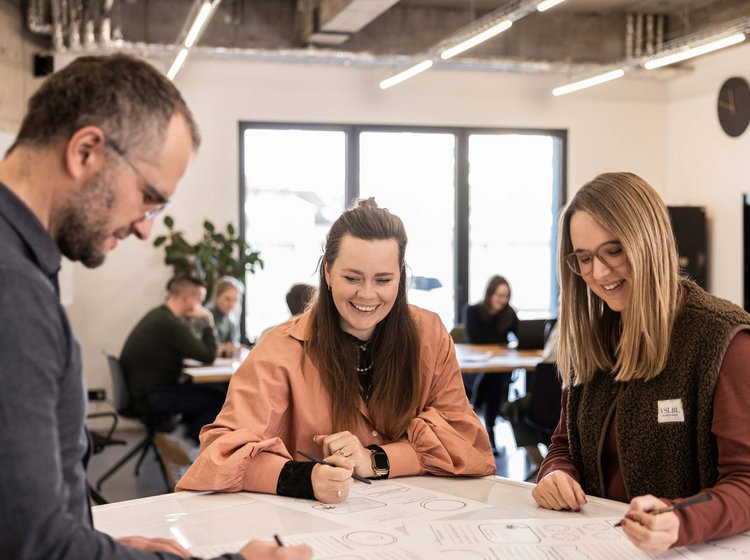 Drei Personen sitzen an einem Tisch und arbeiten gemeinsam an einem großen Plan oder Entwurf. Sie wirken konzentriert und engagiert in einem modernen Büro mit mehreren weiteren Personen im Hintergrund.