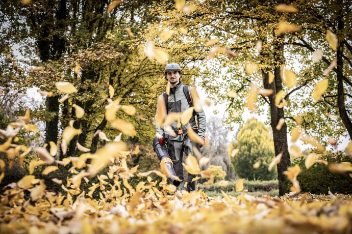 Ein Mann bläst mit einem Laubbläser bunte Herbstblätter auf einem mit Bäumen gesäumten Weg zusammen.