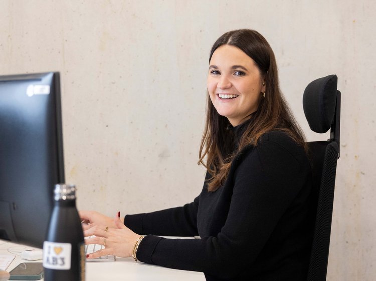 Frau sitzt lächelnd an einem Schreibtisch vor einem Computerbildschirm und arbeitet. Sie trägt schwarze Kleidung und hat braune Haare.