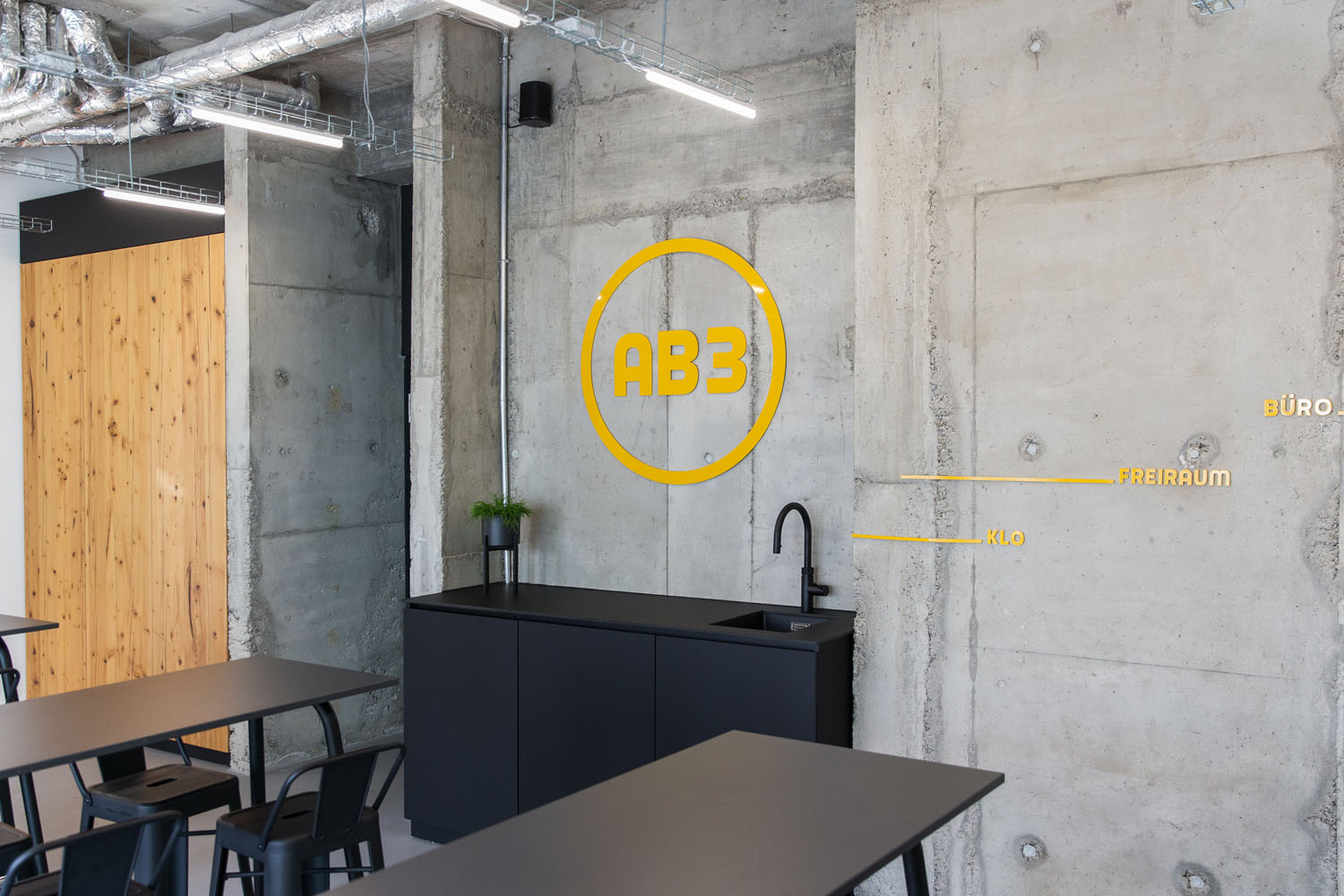 AB3-Logo über Waschbecken Gelbes AB3-Logo auf Betonwand vor einem modernen Tisch mit Stühlen.